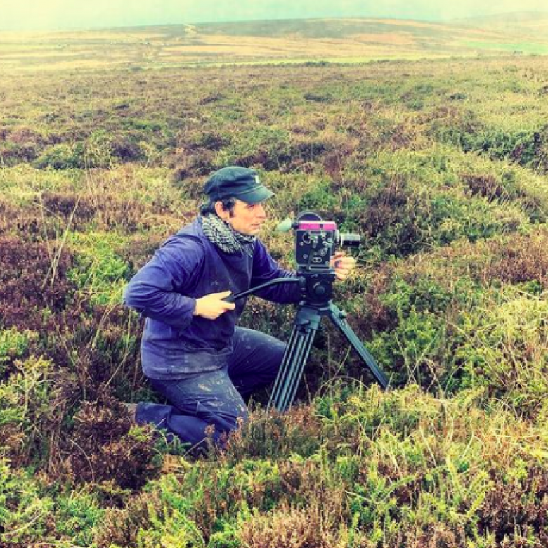 A filmmaker crouches behind his camera amongst shrubbery